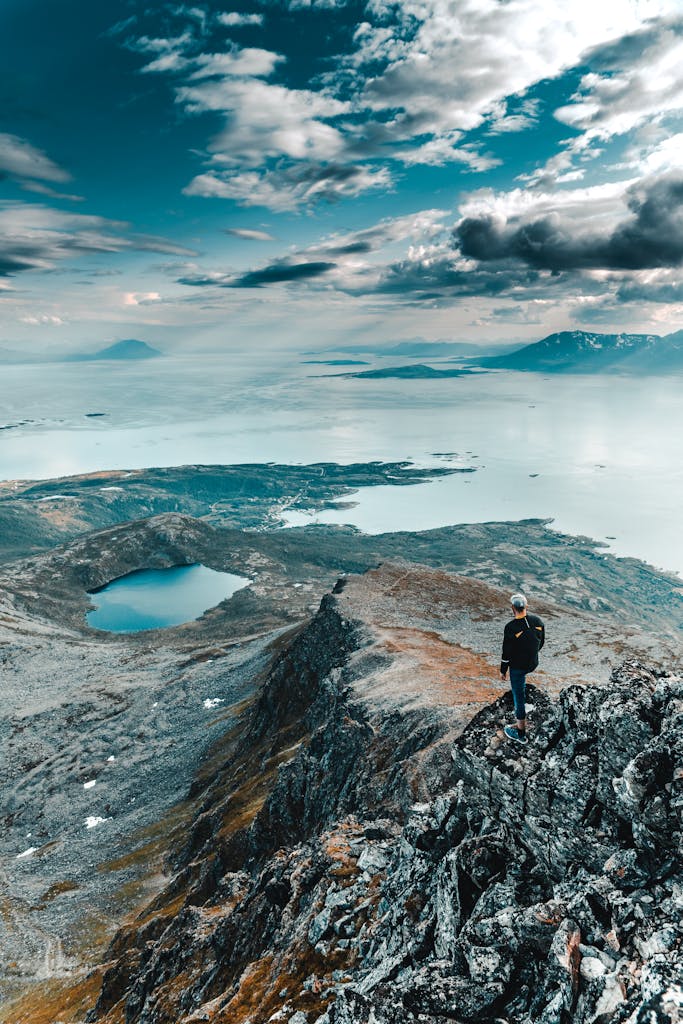 A lone hiker stands on a rugged mountain peak, overlooking a breathtaking landscape under a cloudy sky.